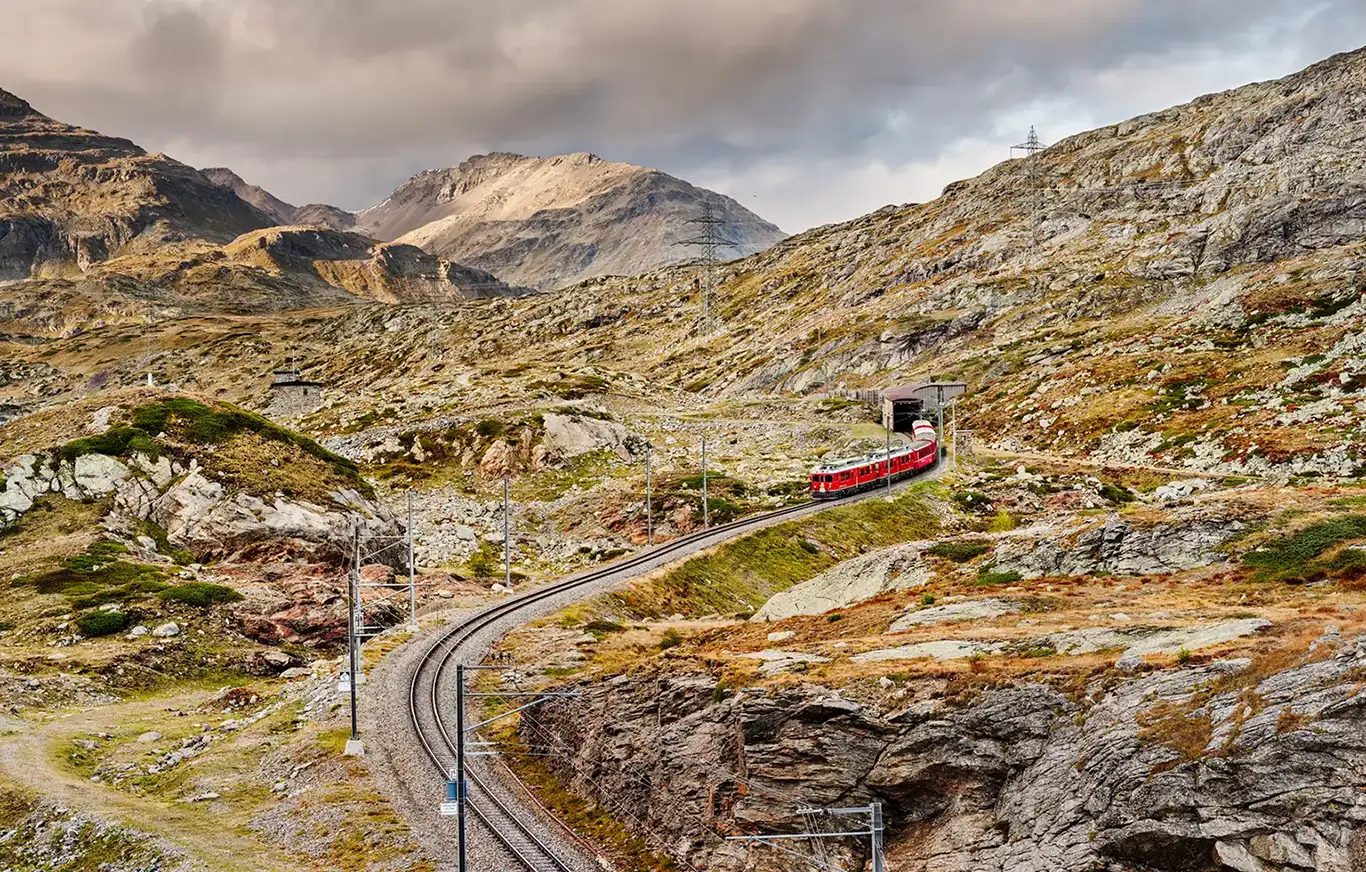 Ein roter Zug der Rhätischen Bahn, angeführt von zwei historischen ABe 4/4 III Triebwagen, schlängelt sich bei letztem Tageslicht durch die alpine Landschaft am Berninapass. Die Schienen führen spektakulär an einem Berghang entlang nahe der Station Alp Grüm. Im Hintergrund erstrecken sich die weiten, rauen Bergketten des UNESCO-Welterbes