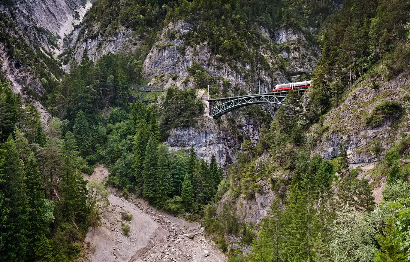 Blick auf die Schlossbachgrabenbrücke, eine eiserne Bogenfachwerkbrücke der Mittenwaldbahn. Die Brücke spannt sich in großer Höhe über eine tiefe, bewaldete und felsige Schlucht. Ein Zug überquert die Brücke vor der Kulisse der schroffen österreichischen Alpen bei klarem Tageslichteichs durch eine der zerklüftetsten Landschaften der Alpen führt